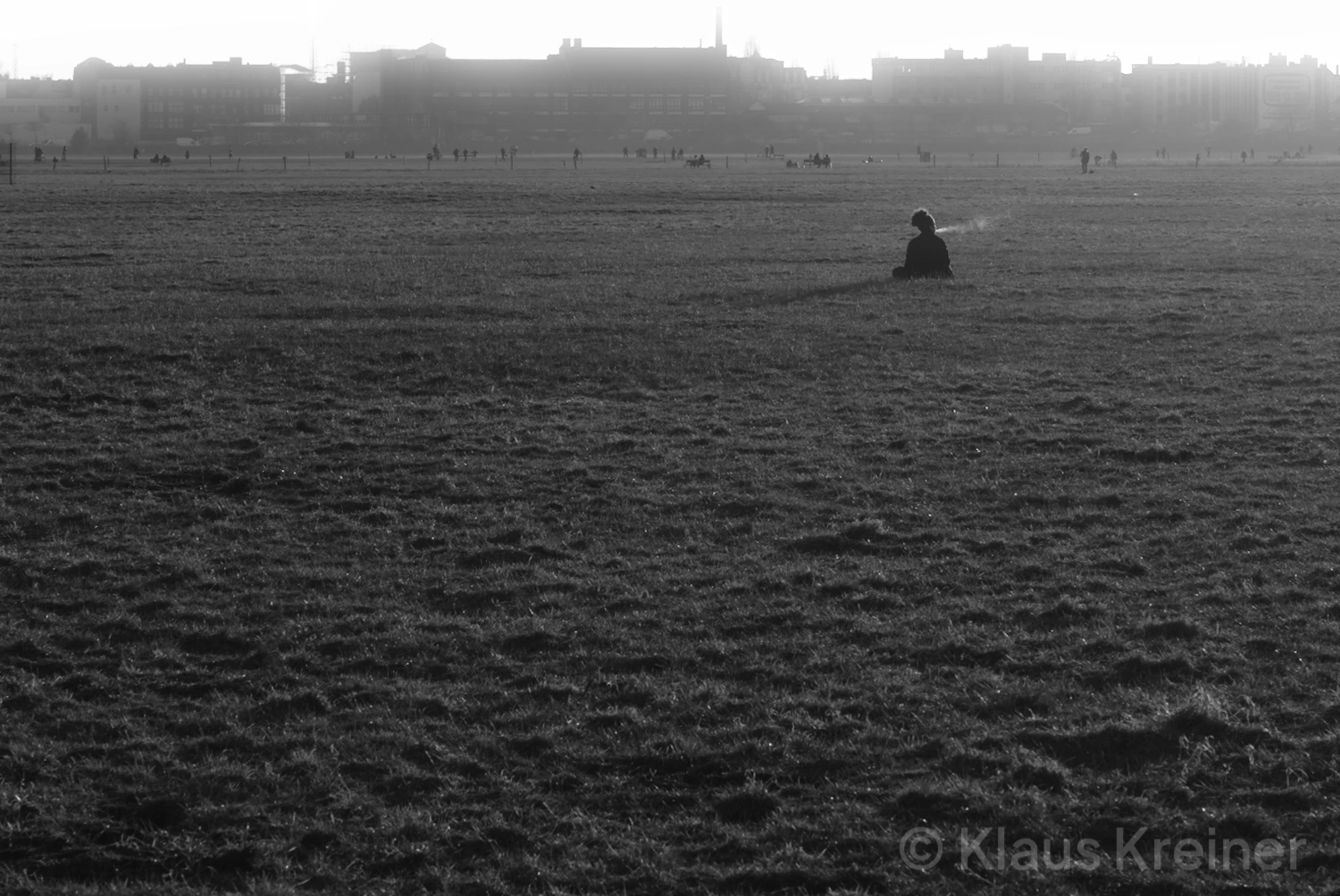 Ende Februar 2021 in Berlin: Ein SW-Foto von einer auf dem Tempelhofer Feld sitzenden, rauchenden Frau, im Horizont sind Menschen.