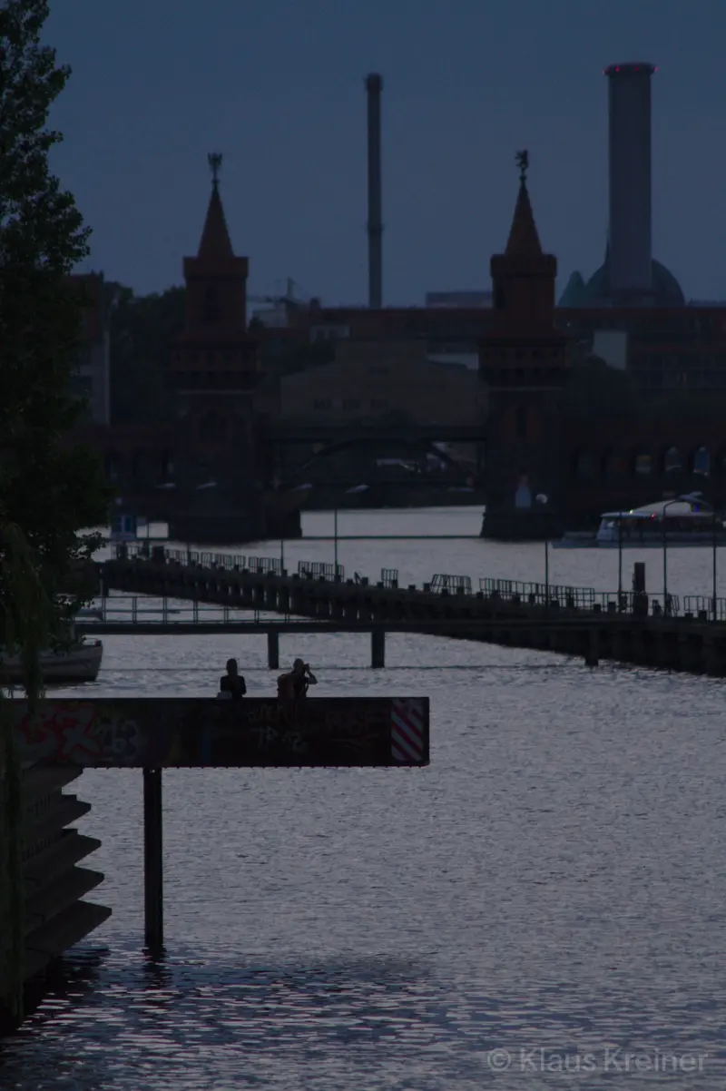 Anfang Juli 2019 in Berlin: Zwei Menschen stehen auf einem weit in das Wasser hineinragenden Steg und schauen zur Oberbaumbrücke.