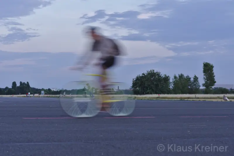 Mitte Juni 2018 in Berlin. Ein Fahrradfahrer wird wegen der Geschwindigkeit verwischt, durch den Rahmen sind sportive Menschen zu sehen.