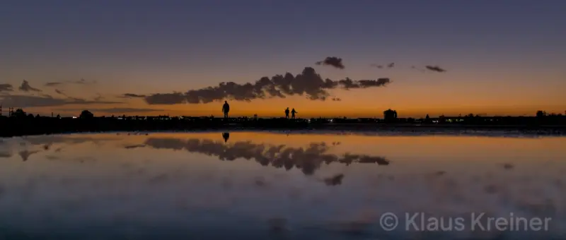 Ende Oktober 2018 in Berlin: Im Spiegelbild einer Wasserfläche geht ein Spaziergänger über das Tempelhofer Feld durch das Abendrot.