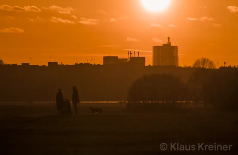 Ende Februar 2022 in Berlin: Ein Pärchen mit Kinderwagen und Hund im Abendrot, im Hintergrund ist die Skyline der Großstadt zu sehen.