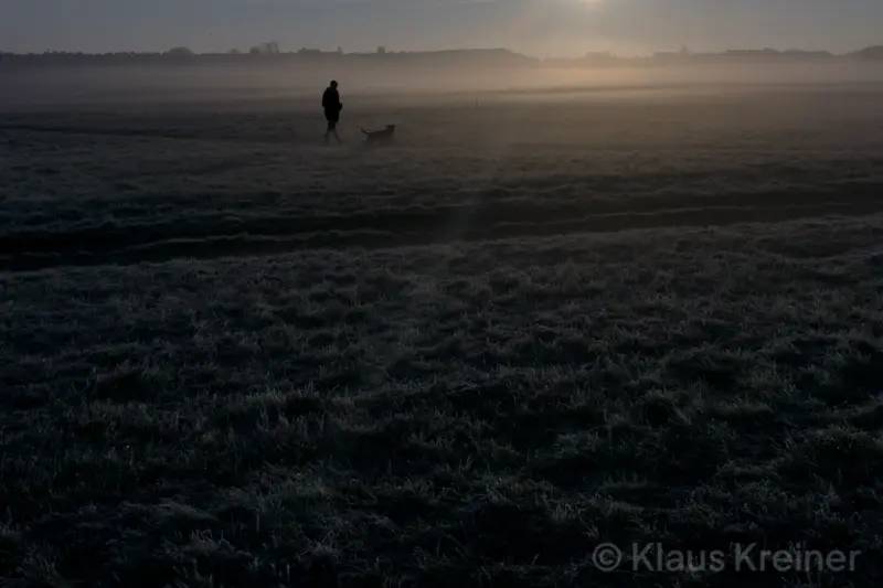Mitte November 2021 in Berlin: Beim Sonnenaufgang geht eine Person mit Hund an einem nebligen Morgen über das Tempelhofer Feld.