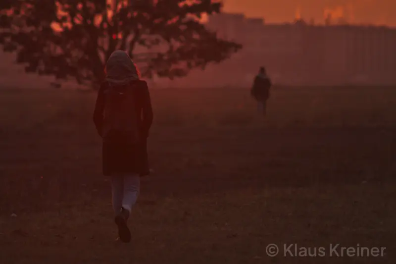 Zwei Personen gehen im Abendrot auf einem Feld an einem Baum vorbei. Im Hintergrund ist eine Skyline einer Stadt zu erkenen.