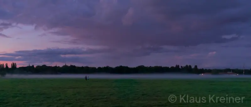 Ein weites Feld mit Abendrotwolken und zwei Menschen nähern sich dem Nebel